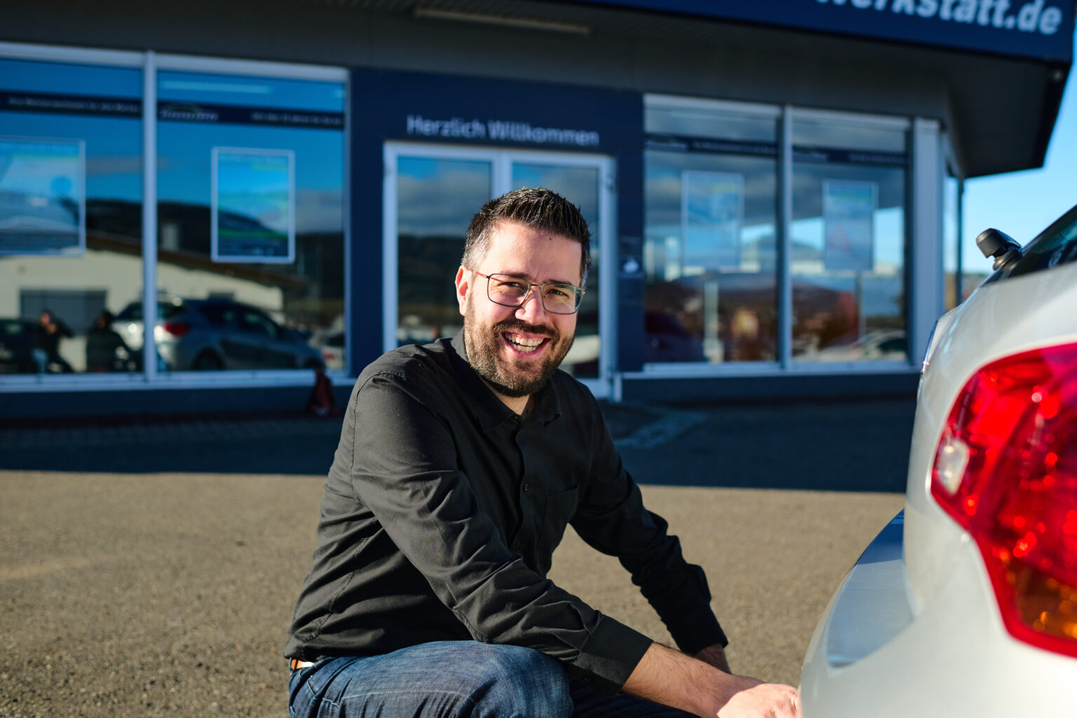 Ein lachender Mann hockt vor dem Heck eines silbernen Autos. Er trägt Jeans, ein schwarzes Oberteil und eine eckige Brille. Mit einer Hand scheint er an die Anhängerkupplung des Wagens zu greifen. Im Hintergrund sieht man unfokussiert die Eingangstür zu einem Gebäude, darüber prangt die Aufschrift "Herzlich Willkommen".